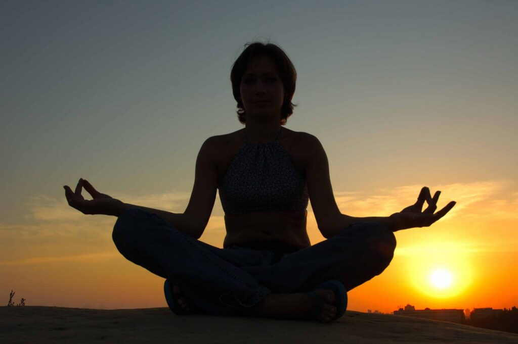 A person is meditating in a peaceful setting with the sun setting in the background.