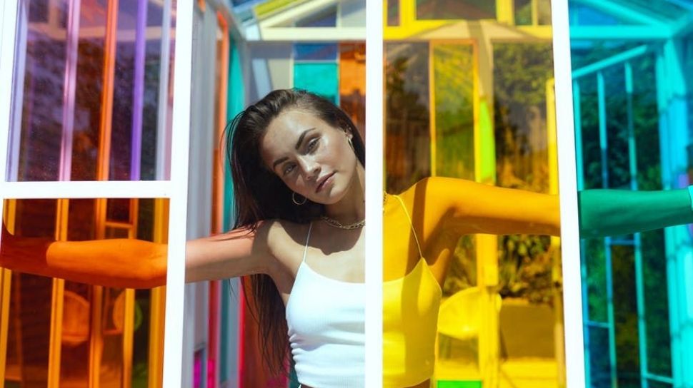 A young woman standing in a colorful glass structure, with her arms outstretched and a confident expression on her face, representing the search query 'Aura rezeki yang terpancar dari diri seseorang'.
