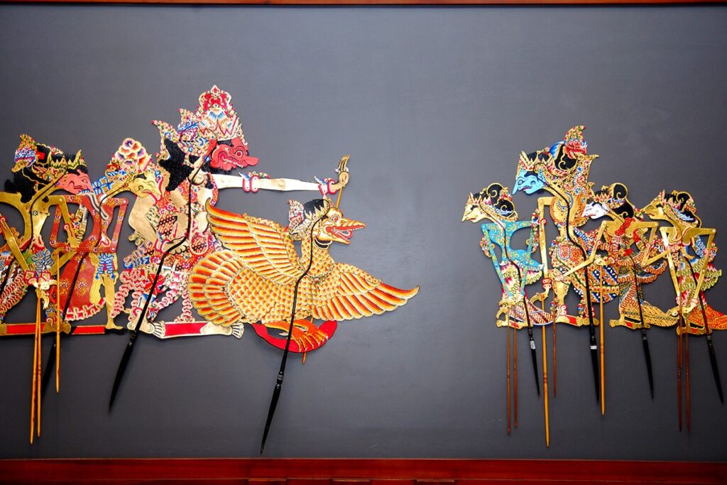 An image of intricately designed Indonesian shadow puppets, made from leather and painted in vivid colors, depicting various negative human traits such as envy, arrogance, greed, revenge, cunning, cruelty, and laziness.