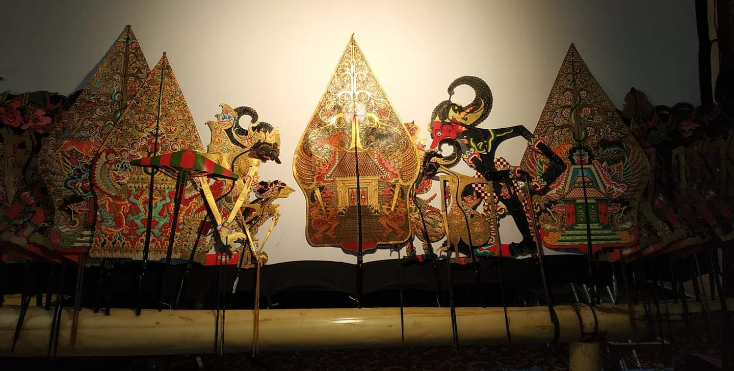 A row of intricately designed and expressive Indonesian Wayang kulit shadow puppets, made from leather and painted with vibrant colors, are displayed against a white background.