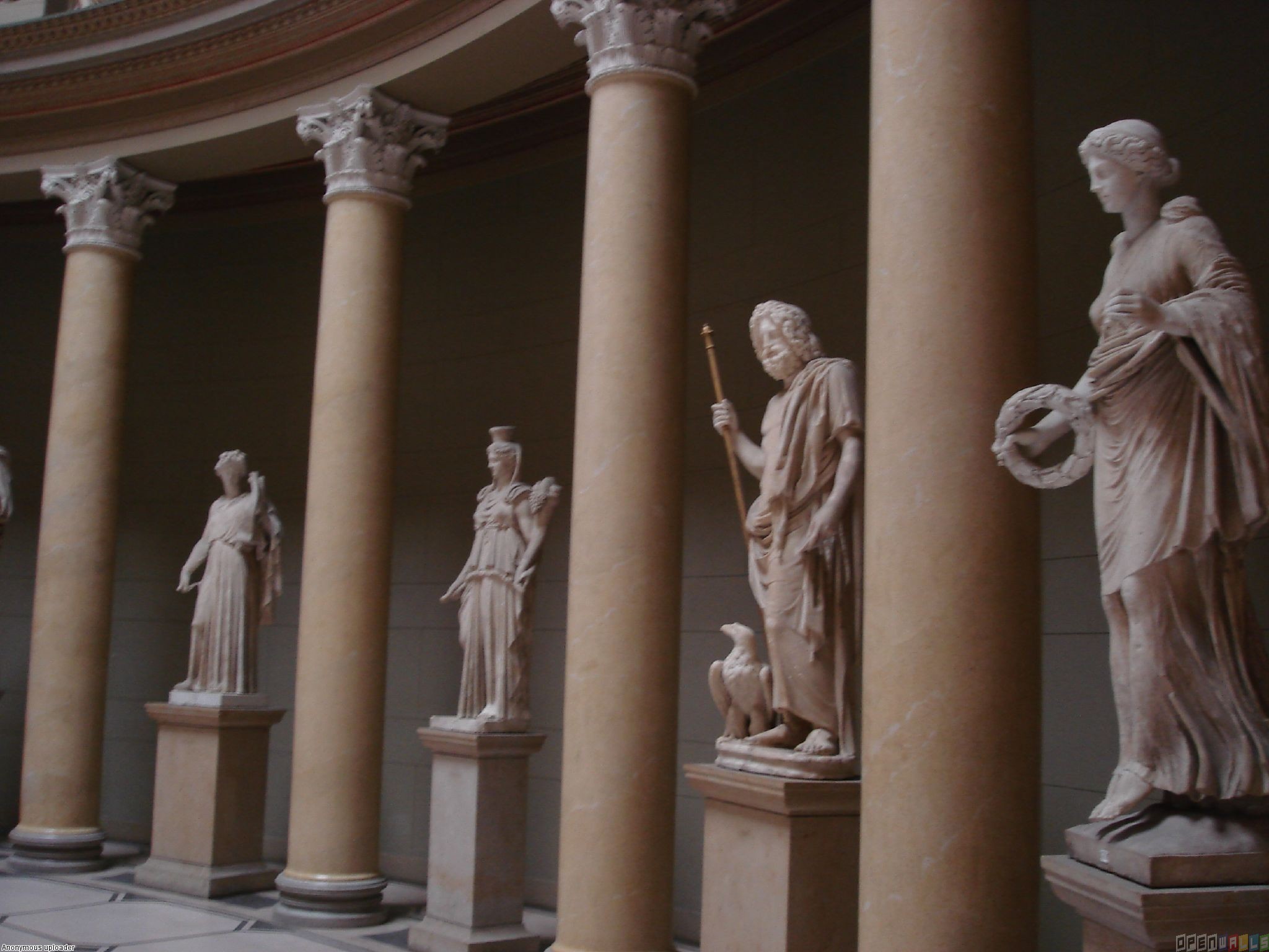 A row of statues of Greek gods and goddesses, including Zeus, Hera, Athena, and Aphrodite, is displayed in a hall with columns.