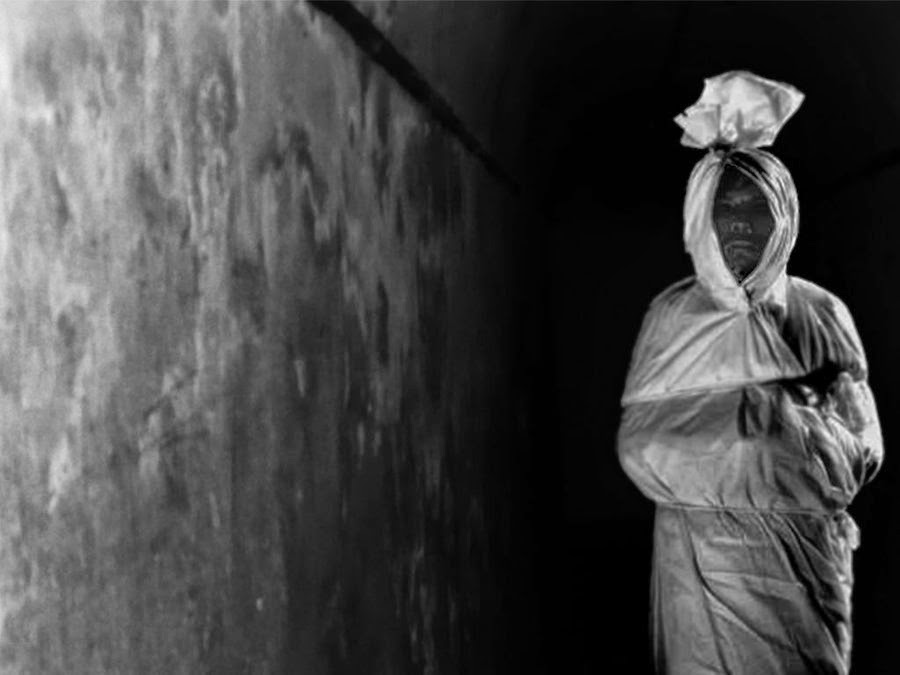 A grayscale image of a pocong ghost, a spirit in Indonesian folklore that is said to be the soul of a deceased person wrapped in a white cloth, standing in a dark hallway.