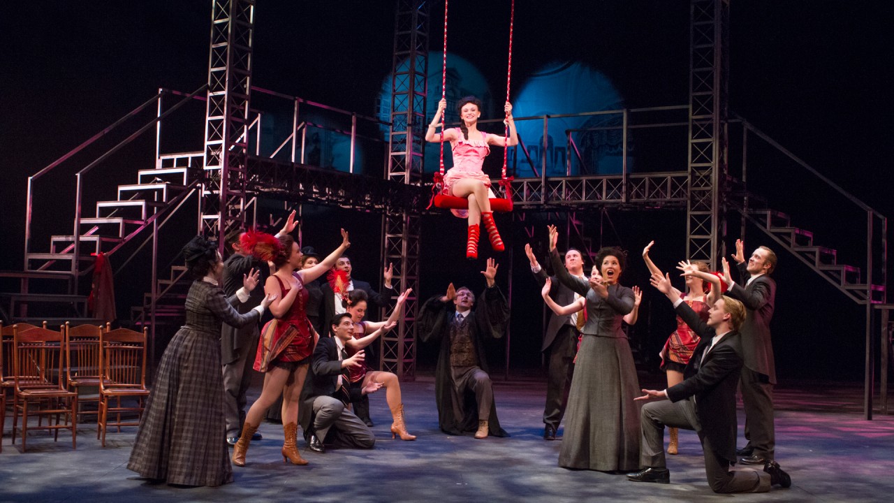 A group of actors perform a folk tale play on stage with one actor swinging on a rope above the rest of the cast.