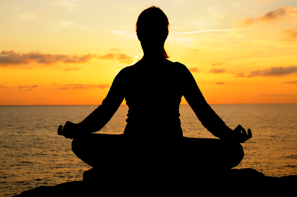A person is meditating on a rock with the ocean behind them and the sky above them is a bright orange as the sun sets.