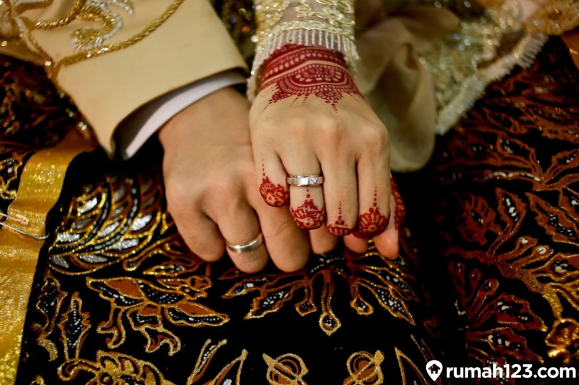 A couple's hands with henna tattoos and wedding rings on top of a traditional Javanese wedding attire, symbolizing their love and commitment, with text 'Nilai weton jodoh Jawa', which means 'The value of Javanese wedding date calculation'.