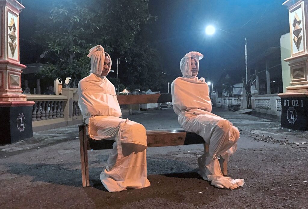 Two people are sitting on a bench at night with white shrouds covering their faces.