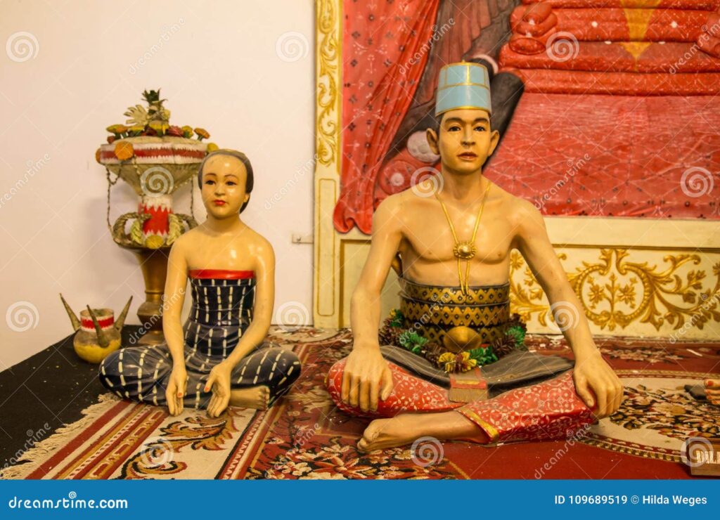 A pair of statues wearing traditional Javanese attire sit on a red and gold patterned carpet, with a red curtain and tapestry with golden patterns in the background.