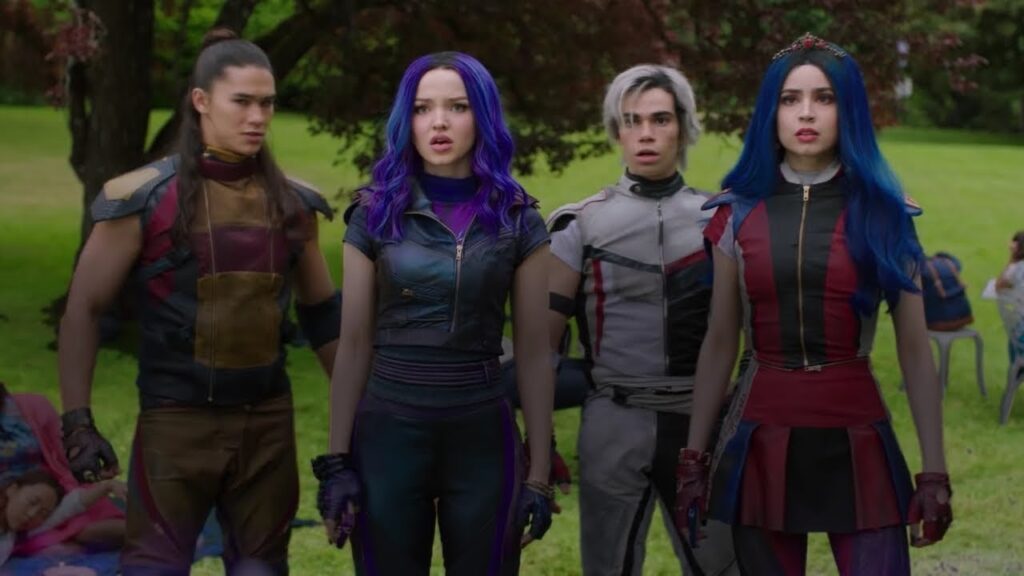The image shows the four main characters from the Disney Channel movie, Descendants, standing in a forest. They are all wearing black and purple clothing. Mal, the daughter of Maleficent, is standing in the center. She has purple hair and is wearing a black leather jacket. Evie, the daughter of the Evil Queen, is standing to the left of Mal. She has blue hair and is wearing a red and black dress. Carlos, the son of Cruella de Vil, is standing to the right of Mal. He has white hair and is wearing a black vest. Jay, the son of Jafar, is standing to the far right. He has black hair and is wearing a black jacket.