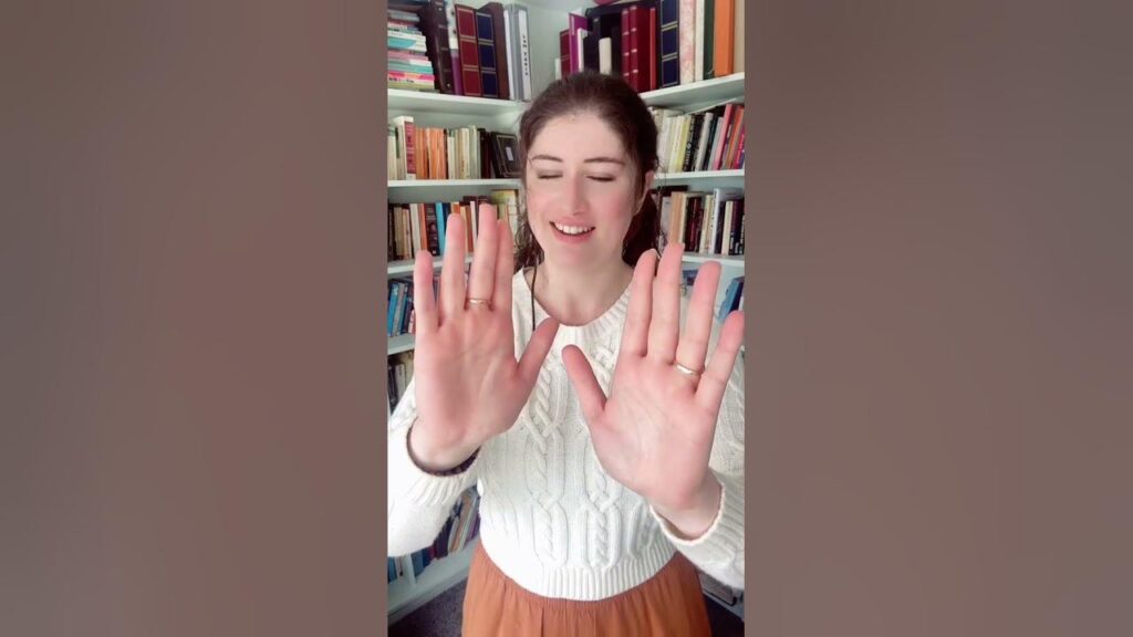A woman in a white sweater and brown skirt demonstrates a technique to restore a closed aura by holding her hands out, palms facing each other.