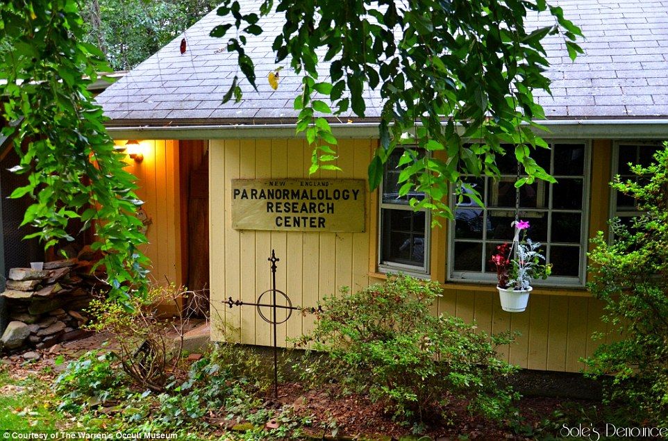 A small, yellow house with a sign that says 'New England Paranormal Research Center' surrounded by overgrown plants and trees.