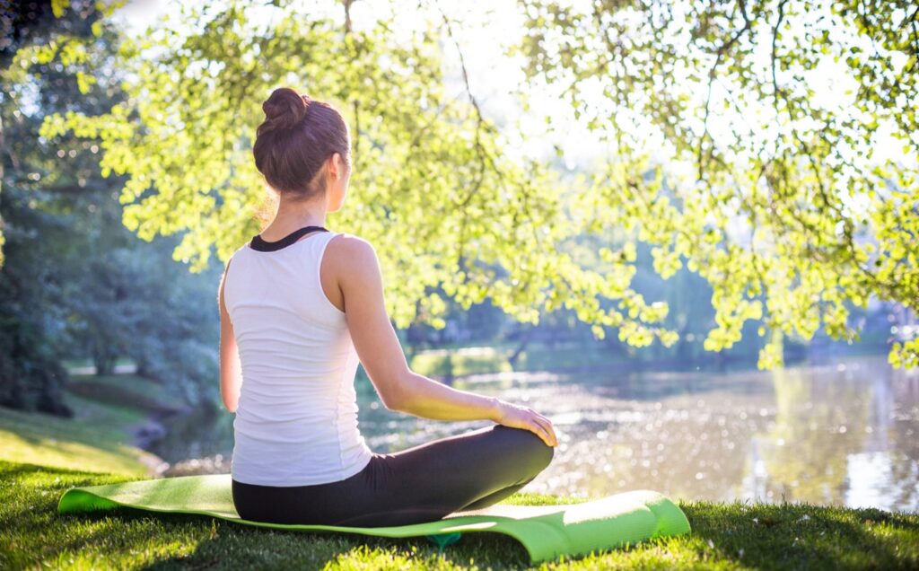 A person sits on a yoga mat in a park, with a lake and trees in the background, and practices meditation and mindfulness.